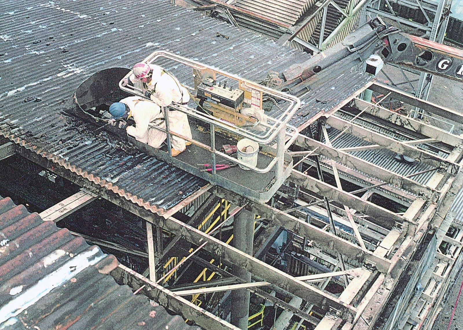 Two workers in coveralls and respirators removing corrugated sheets from a platform